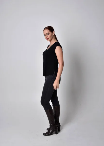 Simple full length portrait of woman with red hair in a ponytail, wearing casual black tshirt and jeans. Standing pose with in side profile, against a  studio background.