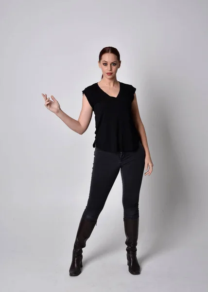 Simple full length portrait of woman with red hair in a ponytail, wearing casual black tshirt and jeans. Standing pose front on with hand gestures, against a  studio background.