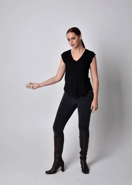 Simple full length portrait of woman with red hair in a ponytail, wearing casual black tshirt and jeans. Standing pose front on with hand gestures, against a  studio background.