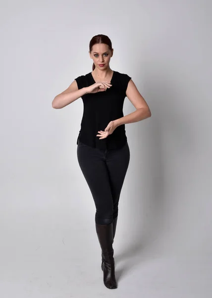 Simple full length portrait of woman with red hair in a ponytail, wearing casual black tshirt and jeans. Standing pose front on with hand gestures, against a  studio background.