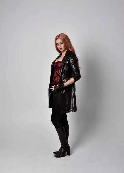 full length portrait of woman with long red hair wearing dark leather coat, corset and boots. Standing pose facing front on wit hand gestures against a  studio background.