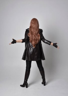 Uzun kızıl saçlı, koyu deri ceketli, korseli ve çizmeli bir kızın tam boy portresi. Stüdyonun arka planında sihirli el hareketleriyle duran poz..