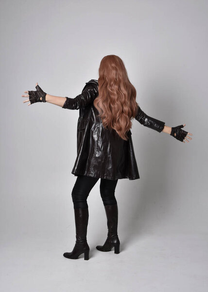 full length portrait of girl with long red hair wearing dark leather coat, corset and boots. Standing pose facing back view with  magical hand gestures against a  studio background.