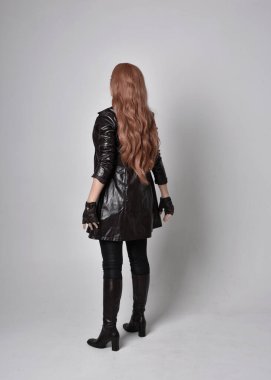 full length portrait of girl with long red hair wearing dark leather coat, corset and boots. Standing pose facing back view with  magical hand gestures against a  studio background. clipart
