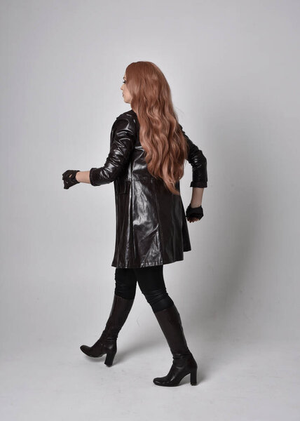 full length portrait of girl with long red hair wearing dark leather coat, corset and boots. Standing pose facing back view with  magical hand gestures against a  studio background.
