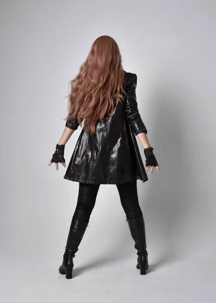 黒い革のコート コルセット ブーツを身に着けている長い赤い髪の少女の完全な長さの肖像画 スタジオの背景に魔法の手のジェスチャーで正面に面した立ちポーズ 低カメラアングル — ストック写真