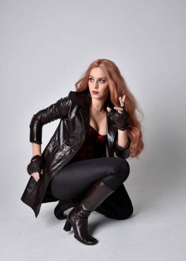 Uzun kızıl saçlı, koyu deri ceketli, korseli ve çizmeli bir kızın tam boy portresi. Oturma pozu, sihirli el hareketleriyle stüdyonun arka planına dönük..