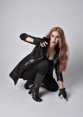 Uzun kızıl saçlı, koyu deri ceketli, korseli ve çizmeli bir kızın tam boy portresi. Oturma pozu, sihirli el hareketleriyle stüdyonun arka planına dönük..