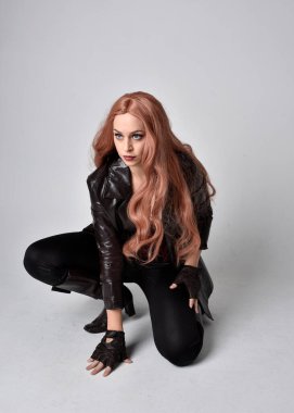 Uzun kızıl saçlı, koyu deri ceketli, korseli ve çizmeli bir kızın tam boy portresi. Oturma pozu, sihirli el hareketleriyle stüdyonun arka planına dönük..