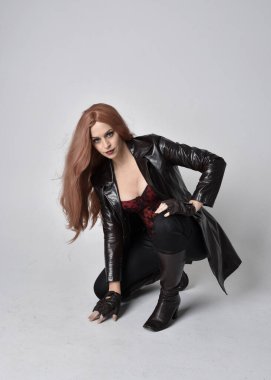 Uzun kızıl saçlı, koyu deri ceketli, korseli ve çizmeli bir kızın tam boy portresi. Oturma pozu, sihirli el hareketleriyle stüdyonun arka planına dönük..