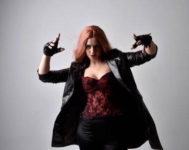 Close up portrait of girl with long red hair wearing dark leather coat, corset and gloves. Posing with gestural hand movements as if casting spell. Moody back lit lighting against a studio background. clipart