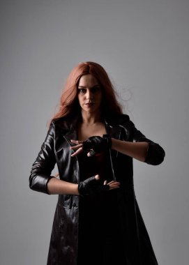 Uzun kızıl saçlı, koyu deri ceketli, korseli ve eldivenli kızın yakın portresi. El hareketleriyle poz vermek, sanki büyü yapmak gibi. Moody arka planda bir stüdyoda ışıklandırma.