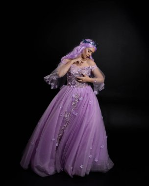Uzun mor fantezi balo elbisesi giyen, uzun boylu, pembe saçlı, stüdyo arka planında zarif hareketlerle duran bir kızın portresi..