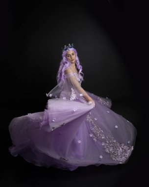 Uzun mor fantezi balo elbisesi giyen, uzun boylu, pembe saçlı, stüdyo arka planında zarif hareketlerle duran bir kızın portresi..