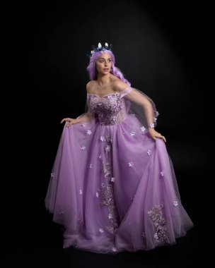 Uzun mor fantezi balo elbisesi giyen, uzun boylu, pembe saçlı, stüdyo arka planında zarif hareketlerle duran bir kızın portresi..