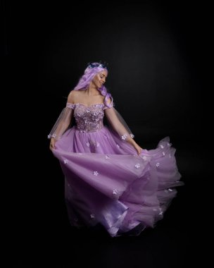 Uzun mor fantezi balo elbisesi giyen, uzun boylu, pembe saçlı, stüdyo arka planında zarif hareketlerle duran bir kızın portresi..
