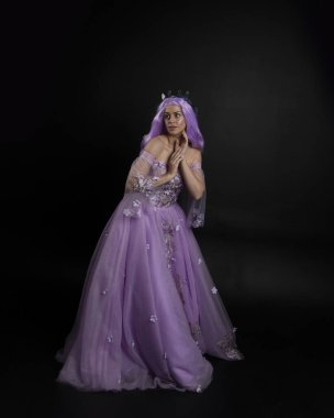 Uzun mor fantezi balo elbisesi giyen, uzun boylu, pembe saçlı, stüdyo arka planında zarif hareketlerle duran bir kızın portresi..