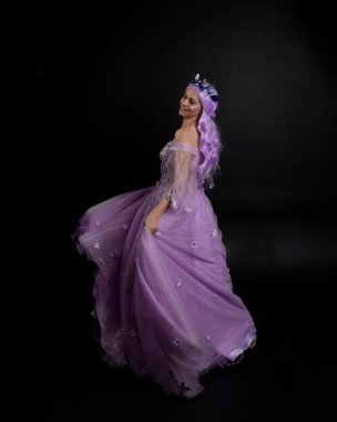 Uzun mor fantezi balo elbisesi giyen, uzun boylu bir kızın portresi. Taçlı ve pembe saçlı. Stüdyo arka planında kameraya dönük poz veriyor..
