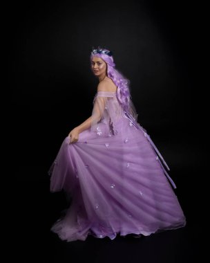 Uzun mor fantezi balo elbisesi giyen, uzun boylu bir kızın portresi. Taçlı ve pembe saçlı. Stüdyo arka planında kameraya dönük poz veriyor..