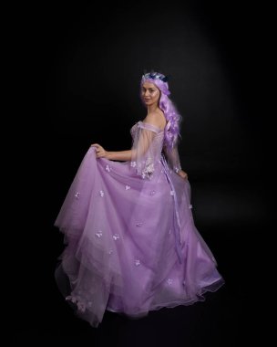 Uzun mor fantezi balo elbisesi giyen, uzun boylu bir kızın portresi. Taçlı ve pembe saçlı. Stüdyo arka planında kameraya dönük poz veriyor..