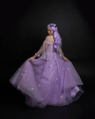 Uzun mor fantezi balo elbisesi giyen, uzun boylu bir kızın portresi. Taçlı ve pembe saçlı. Stüdyo arka planında kameraya dönük poz veriyor..