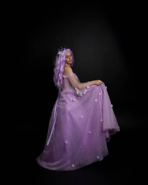 Uzun mor fantezi balo elbisesi giyen, uzun boylu bir kızın portresi. Taçlı ve pembe saçlı. Stüdyo arka planında kameraya dönük poz veriyor..