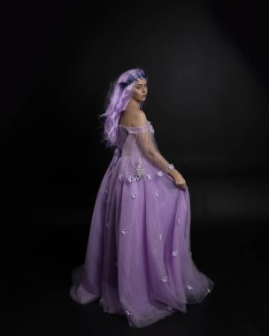 Uzun mor fantezi balo elbisesi giyen, uzun boylu bir kızın portresi. Taçlı ve pembe saçlı. Stüdyo arka planında kameraya dönük poz veriyor..