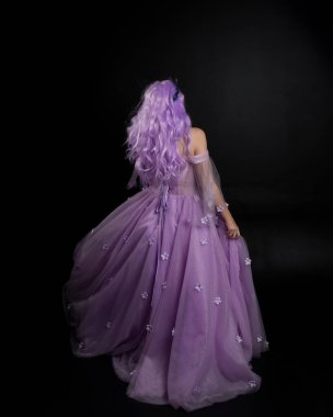 Uzun mor fantezi balo elbisesi giyen, uzun boylu bir kızın portresi. Taçlı ve pembe saçlı. Stüdyo arka planında kameraya dönük poz veriyor..