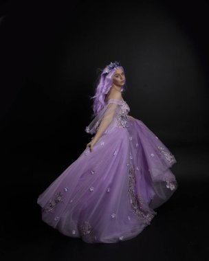 Uzun mor fantezi balo elbisesi giyen, uzun boylu bir kızın portresi. Taçlı ve pembe saçlı. Stüdyo arka planında kameraya dönük poz veriyor..