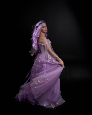 Uzun mor fantezi balo elbisesi giyen, uzun boylu bir kızın portresi. Taçlı ve pembe saçlı. Stüdyo arka planında kameraya dönük poz veriyor..