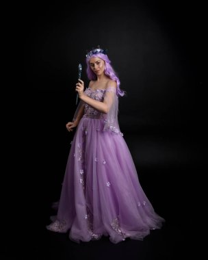 Uzun mor fantezi balo elbisesi giyen, uzun boylu bir kızın portresi. Taçlı ve pembe saçlı. Stüdyo arka planında değnek tutan bir duruş..