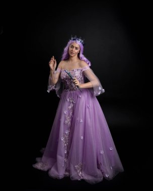 Uzun mor fantezi balo elbisesi giyen, uzun boylu bir kızın portresi. Taçlı ve pembe saçlı. Stüdyo arka planında değnek tutan bir duruş..