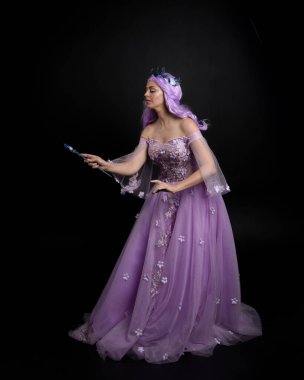 Uzun mor fantezi balo elbisesi giyen, uzun boylu bir kızın portresi. Taçlı ve pembe saçlı. Stüdyo arka planında değnek tutan bir duruş..