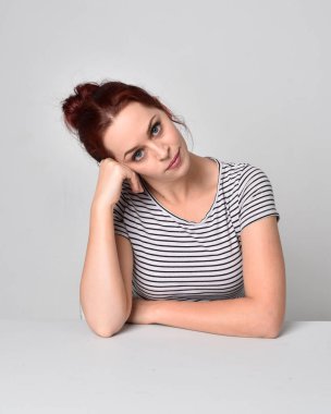 Kırmızı saçlı, çizgili gömlek giyen bir kızın 3 / 4 'lük yakın portresi. Stüdyo arka planında masa pozuna yaslan.