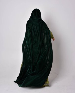 Fantezi kadife pelerinli yeşil ortaçağ elbisesi giyen kızıl saçlı kızın tam boy portresi. Stüdyonun arka planında yalnız poz vermek.