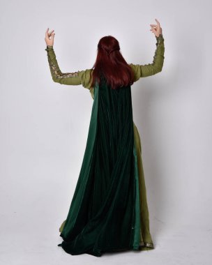 Fantezi kadife pelerinli yeşil ortaçağ elbisesi giyen kızıl saçlı kızın tam boy portresi. Stüdyonun arka planında yalnız poz vermek.