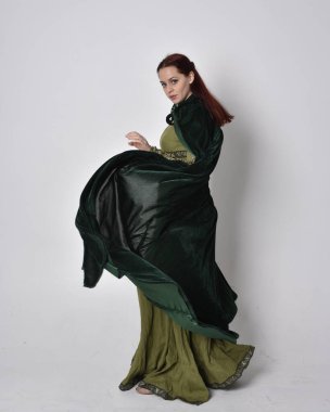 Fantezi kadife pelerinli yeşil ortaçağ elbisesi giyen kızıl saçlı kızın tam boy portresi. Stüdyonun arka planında yalnız poz vermek.