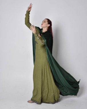 Fantezi kadife pelerinli yeşil ortaçağ elbisesi giyen kızıl saçlı kızın tam boy portresi. Stüdyonun arka planında yalnız poz vermek.