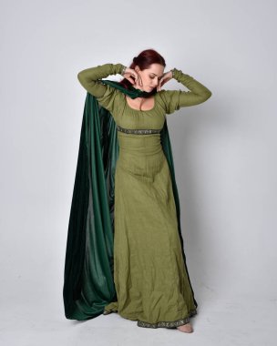 Fantezi kadife pelerinli yeşil ortaçağ elbisesi giyen kızıl saçlı kızın tam boy portresi. Stüdyonun arka planında yalnız poz vermek.