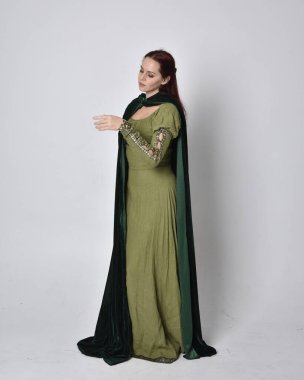 Fantezi kadife pelerinli yeşil ortaçağ elbisesi giyen kızıl saçlı kızın tam boy portresi. Stüdyonun arka planında yalnız poz vermek.