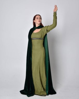 Fantezi kadife pelerinli yeşil ortaçağ elbisesi giyen kızıl saçlı kızın tam boy portresi. Stüdyonun arka planında yalnız poz vermek.