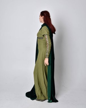 Fantezi kadife pelerinli yeşil ortaçağ elbisesi giyen kızıl saçlı kızın tam boy portresi. Stüdyonun arka planında yalnız poz vermek.