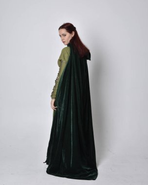 Fantezi kadife pelerinli yeşil ortaçağ elbisesi giyen kızıl saçlı kızın tam boy portresi. Stüdyonun arka planında yalnız poz vermek.