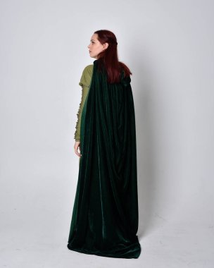 Fantezi kadife pelerinli yeşil ortaçağ elbisesi giyen kızıl saçlı kızın tam boy portresi. Stüdyonun arka planında yalnız poz vermek.