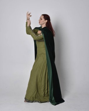 Fantezi kadife pelerinli yeşil ortaçağ elbisesi giyen kızıl saçlı kızın tam boy portresi. Stüdyonun arka planında yalnız poz vermek.