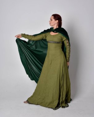 Fantezi kadife pelerinli yeşil ortaçağ elbisesi giyen kızıl saçlı kızın tam boy portresi. Stüdyonun arka planında yalnız poz vermek.