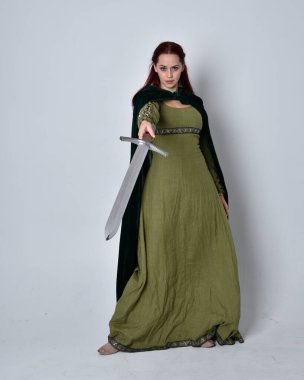 Fantezi kadife pelerinli yeşil ortaçağ elbisesi giyen kızıl saçlı kızın tam boy portresi. Stüdyonun arka planında yalnız poz vermek.