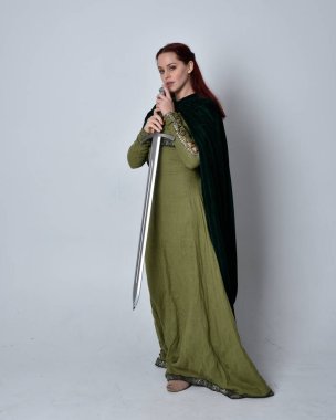 Fantezi kadife pelerinli yeşil ortaçağ elbisesi giyen kızıl saçlı kızın tam boy portresi. Stüdyonun arka planında yalnız poz vermek.