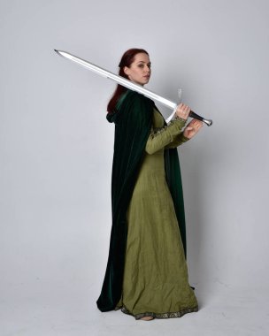 Fantezi kadife pelerinli yeşil ortaçağ elbisesi giyen kızıl saçlı kızın tam boy portresi. Stüdyonun arka planında yalnız poz vermek.