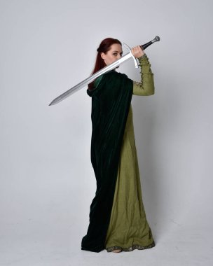 Fantezi kadife pelerinli yeşil ortaçağ elbisesi giyen kızıl saçlı kızın tam boy portresi. Stüdyonun arka planında yalnız poz vermek.